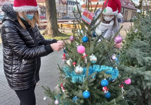 Pracownicy w czapkach Mikołajowych zawieszają bombki na choince